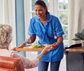 Female Care Worker In Uniform Bringing Meal On Tray To Senior Wo Female Care Worker In Uniform Bringing Meal On Tray To Senior Woman Sitting In Lounge At Home